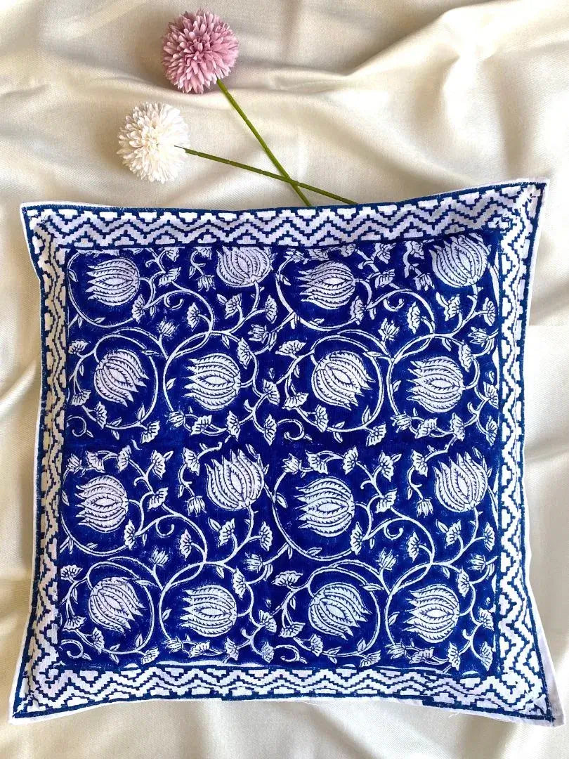 A hand blockprinted cushion cover in blue with a lotus design, displayed on a white surface, accompanied by a floral decoration.
